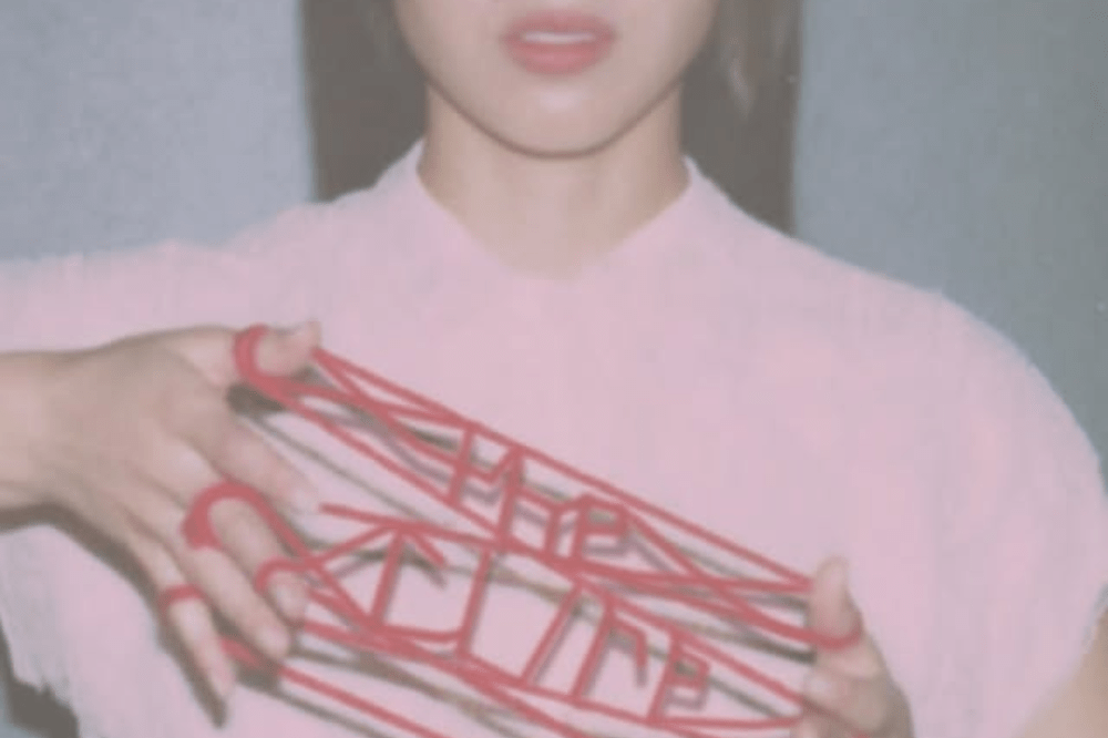 Pessoa veste blusa rosa enquanto segura fios vermelhos entre os dedos, formando um desenho geométrico e o texto "The Cure" diante da câmera