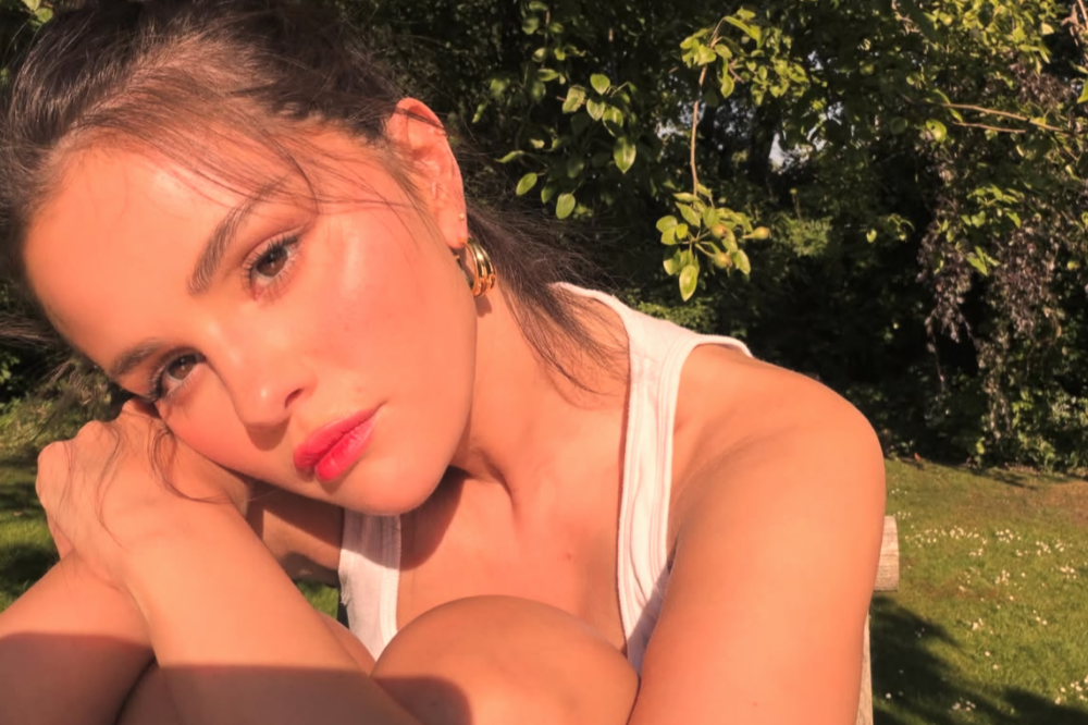Selena Gomez com expressão séria, rosto iluminado pelo sol, usando batom rosa e brincos de argola dourados, em um jardim verde