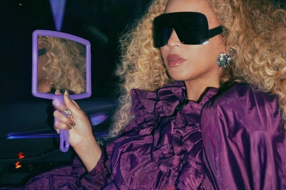 Beyoncé, com cabelo loiro cacheado, usando óculos escuros grandes, batom rosa brilhante, brinco prateado e blusa roxa com babados, segura um espelho roxo que reflete seu cabelo