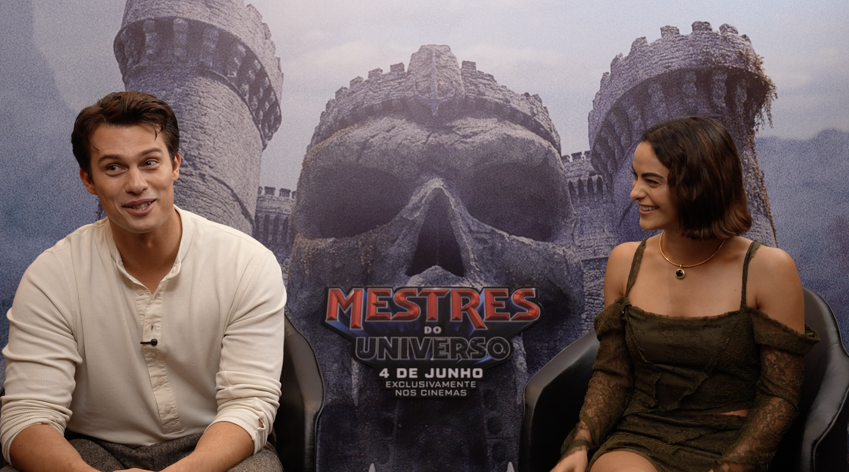 Um homem e uma mulher jovens sorriem sentados, com um banner do Castelo de Grayskull e o título "Mestres do Universo" ao fundo