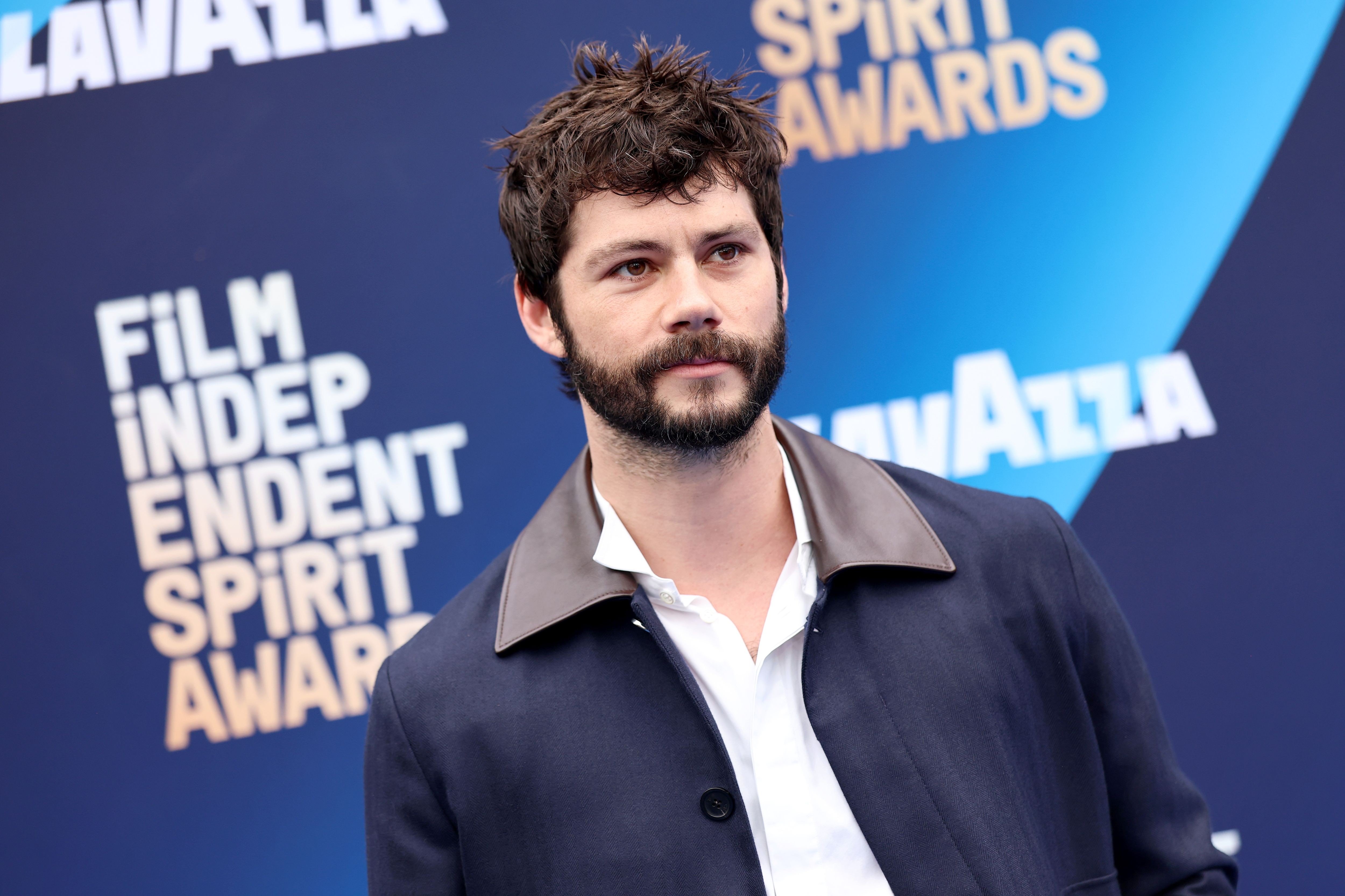 Dylan O'Brien, ator com barba e cabelo castanhos bagunçados, veste camisa branca e jaqueta azul-marinho com gola marrom, posando em frente a um painel azul com os logos "Film Independent Spirit Awards" e "Lavazza"