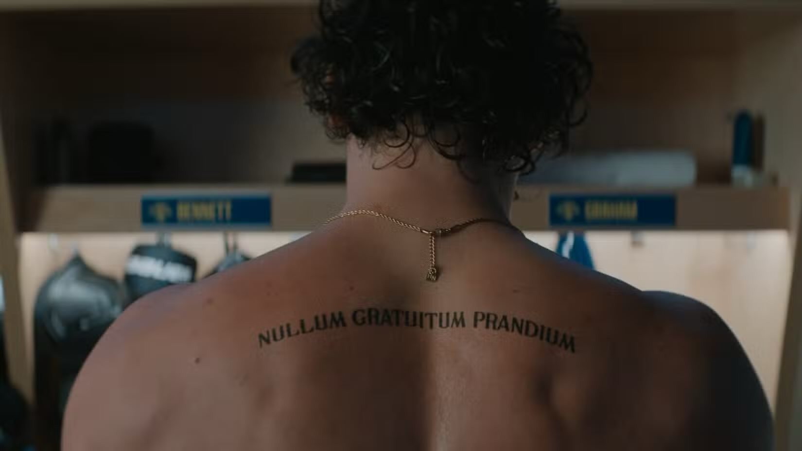 Costas de um homem musculoso com cabelo cacheado escuro, usando um colar dourado e uma tatuagem em latim 