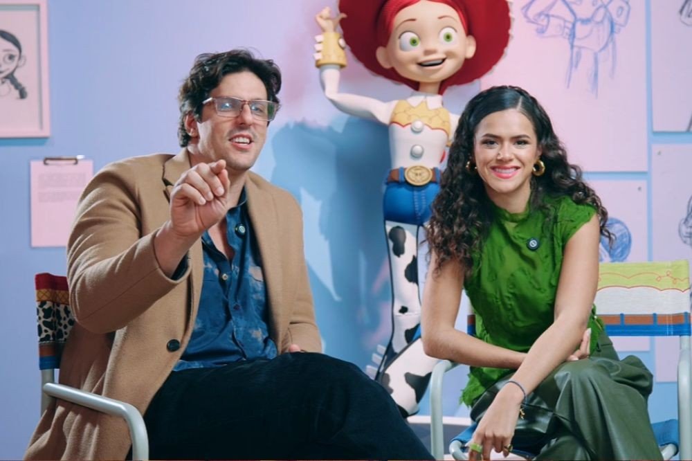 Um homem e uma mulher sentados em cadeiras de diretor, com uma estátua da personagem Jessie de Toy Story ao fundo. O homem, à esquerda, usa óculos, camisa azul e casaco bege, gesticulando com a mão direita. A mulher, à direita, tem cabelos cacheados, veste blusa verde e calça verde-escura, sorrindo para a câmera. Desenhos de personagens de animação decoram a parede azul clara atrás deles