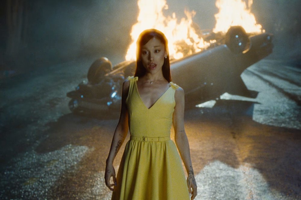 Ariana Grande, com cabelo castanho solto e vestido amarelo, olha para frente com expressão séria, enquanto um carro capotado queima ao fundo em uma rua escura e molhada