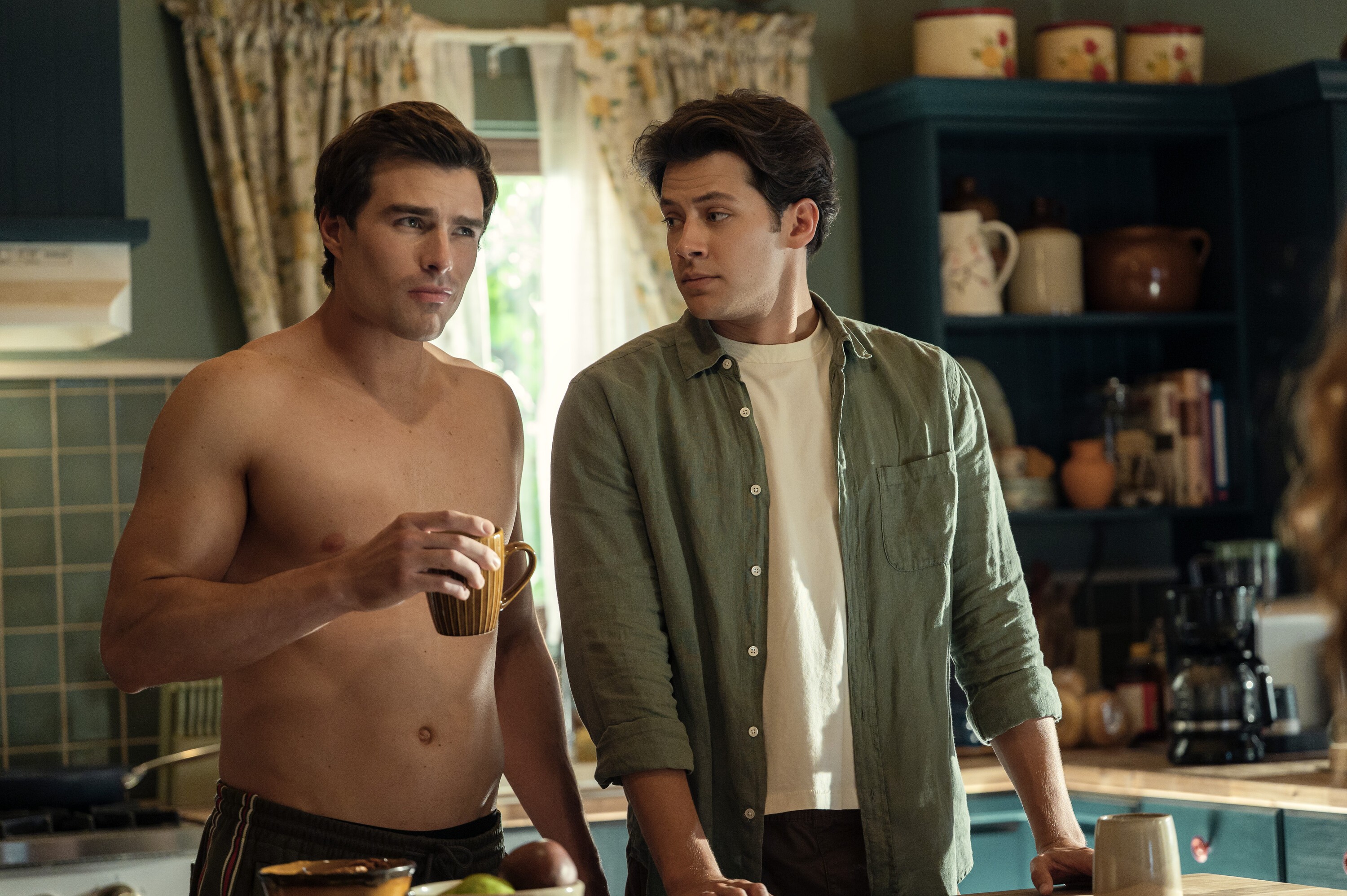Dois homens jovens na cozinha. O da esquerda, sem camisa e com shorts escuros, segura uma caneca marrom. O da direita, com camisa verde-oliva e camiseta branca, apoia-se na bancada, ambos olham para a direita.