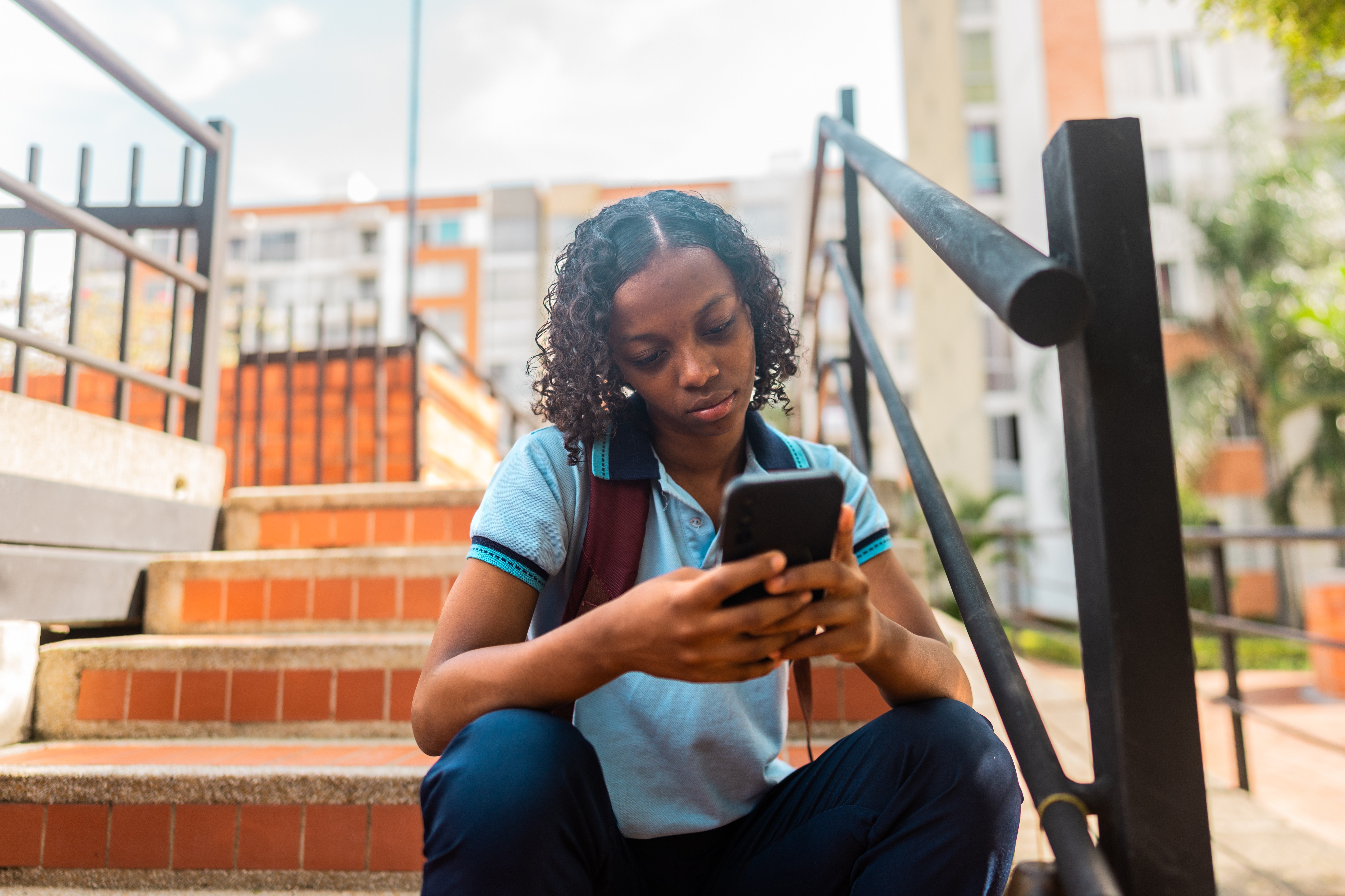 Jovem negra com cabelo cacheado, vestindo uniforme escolar azul claro e calça escura, sentada em escadas de tijolos, concentrada no celular. Ela usa uma mochila marrom e está ao ar livre, com prédios ao fundo