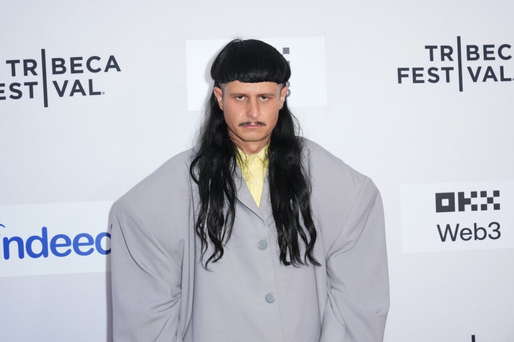 Homem com cabelo preto liso, corte tigela na frente e longo atrás, bigode fino, vestindo camisa amarela e paletó cinza oversized, em frente a um painel branco com logos do Tribeca Festival, Indeed e OKX Web3