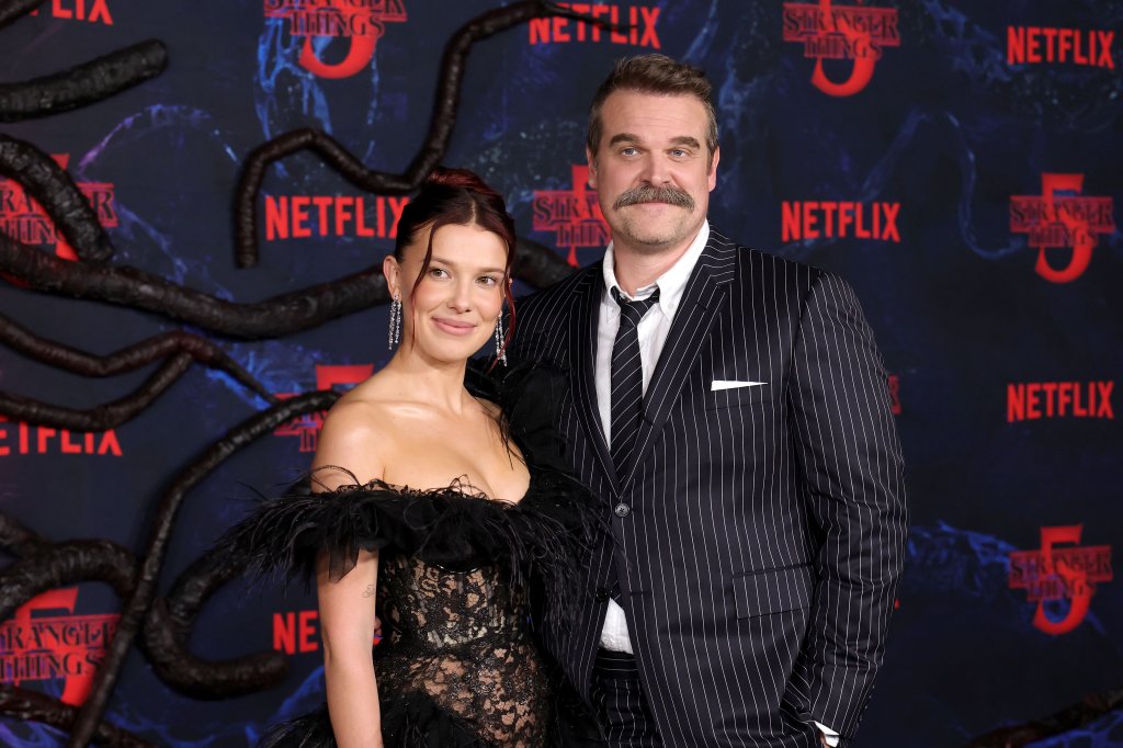 Millie Bobby Brown e David Harbour sorriem para a câmera em um evento. Millie usa um vestido preto de renda com detalhes de penas e brincos longos. David veste um terno risca de giz com camisa branca e gravata preta, ostentando um bigode. Ao fundo, um painel escuro com o logo Stranger Things 5 e tentáculos escuros