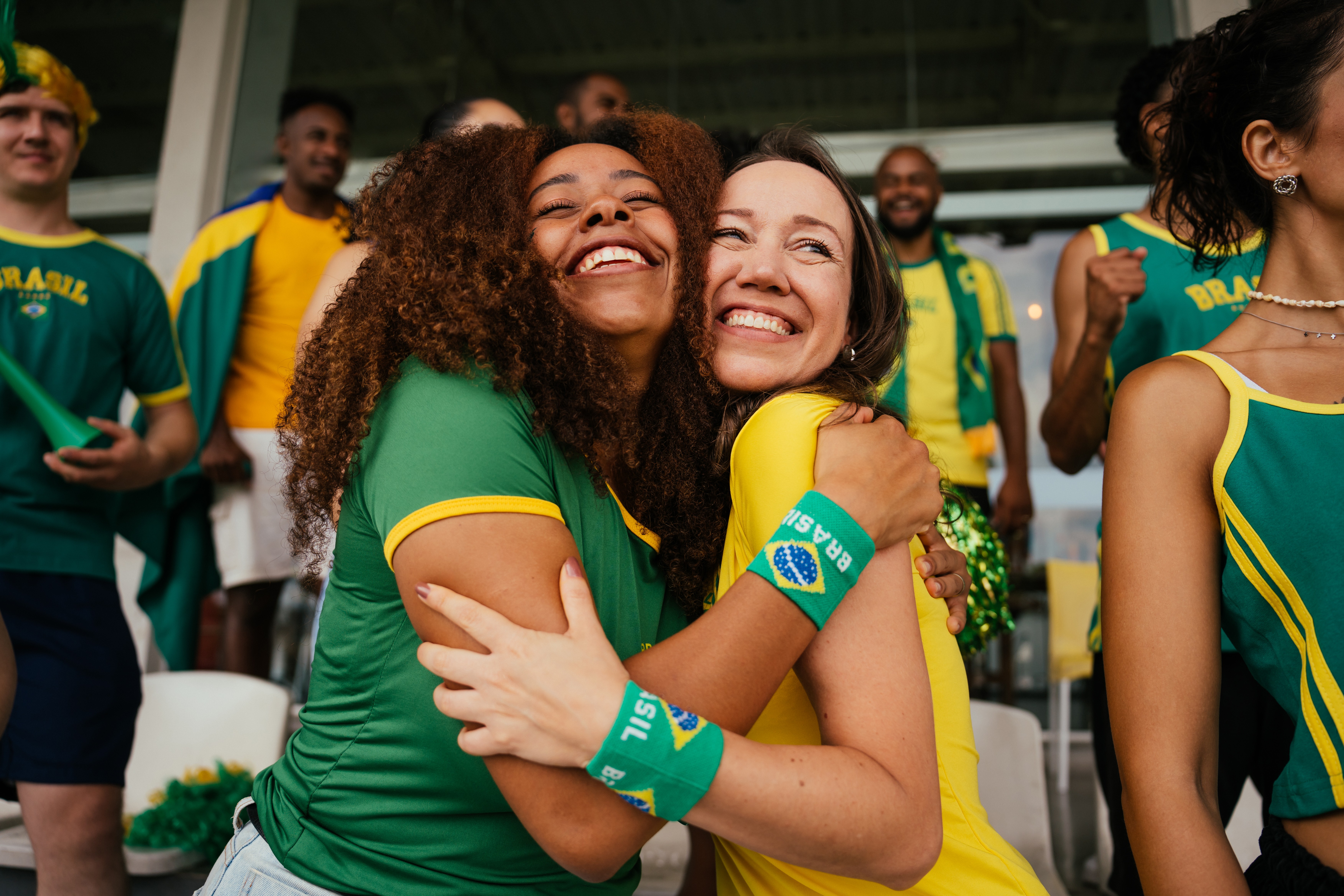 Duas mulheres sorridentes, uma com cabelo cacheado e camisa verde, outra com camisa amarela e munhequeira do Brasil, se abraçam em uma arquibancada cheia de torcedores brasileiros, celebrando com alegria
