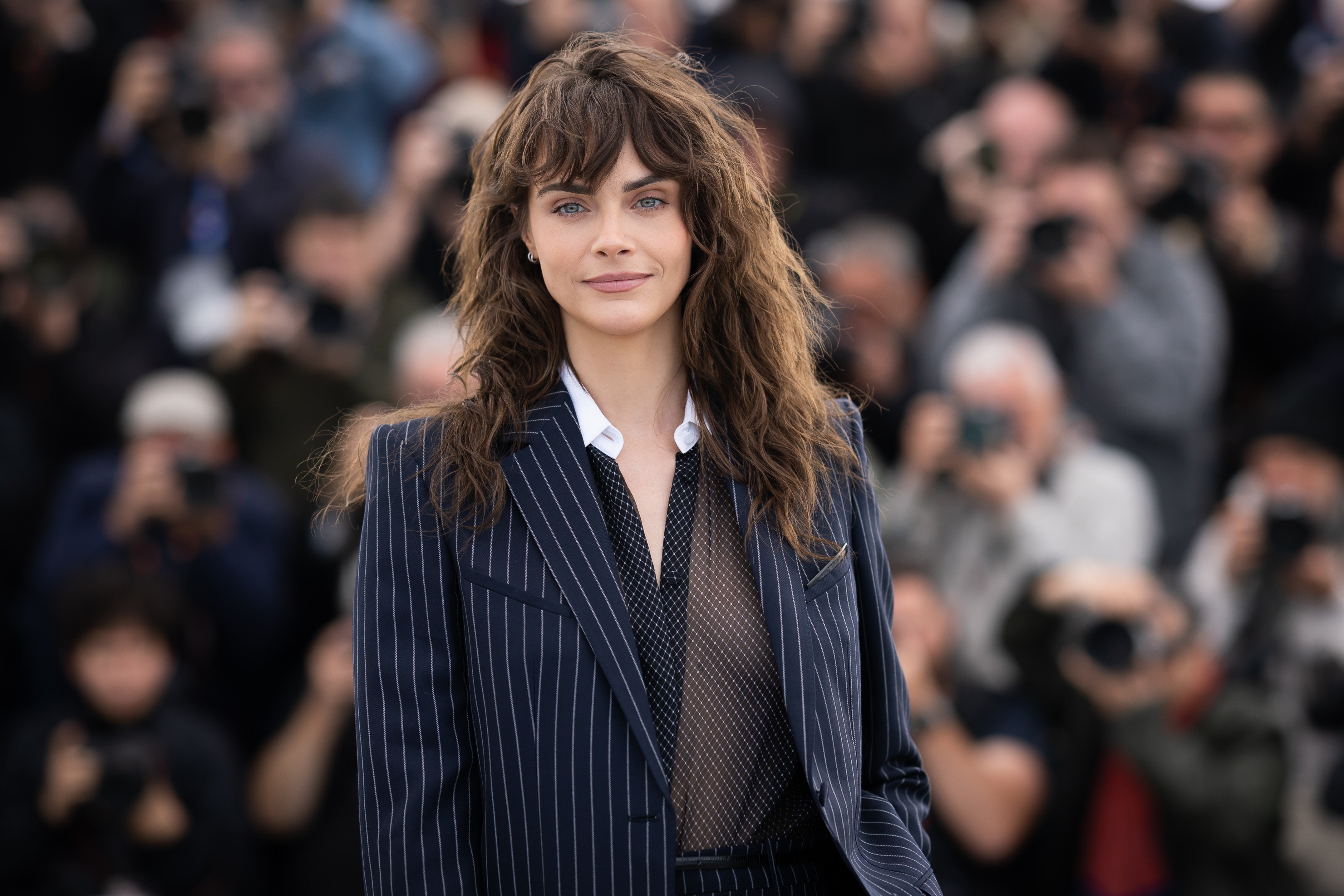 Mulher de cabelos castanhos ondulados e franja, usando blazer azul-marinho listrado, camisa branca e blusa preta transparente, sorri levemente para a câmera. Ao fundo, uma multidão de fotógrafos desfocados.