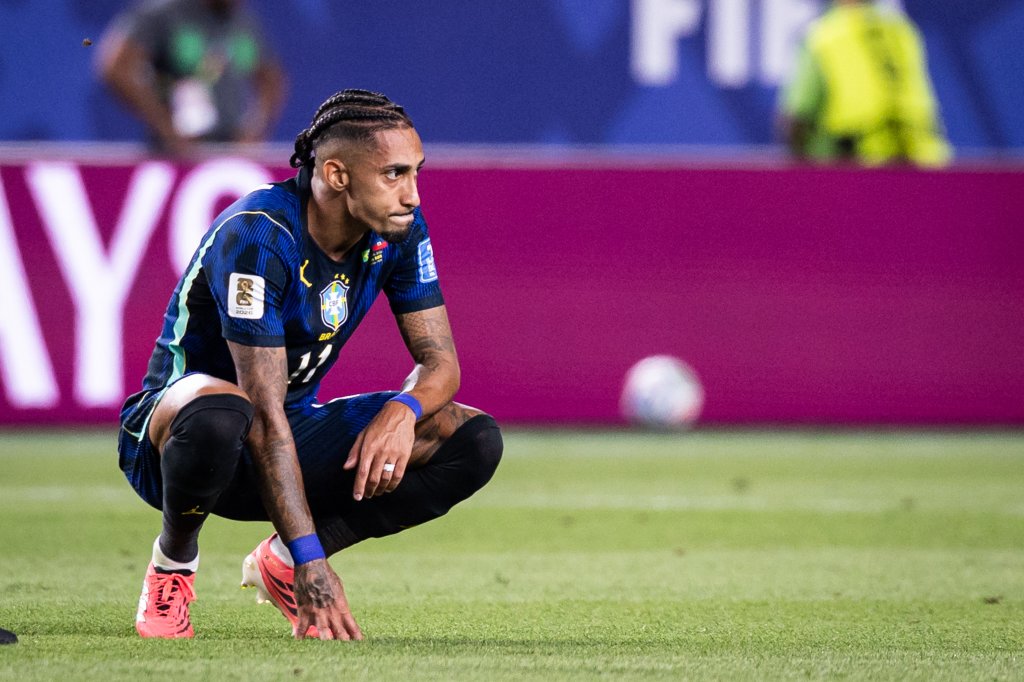 Jogador de futebol com tranças, vestindo uniforme azul da seleção brasileira, agachado no campo com expressão de desânimo, olhando para o lado direito. Ele usa chuteiras rosa e uma braçadeira azul no pulso esquerdo. Ao fundo, uma barreira magenta e um campo de futebol.