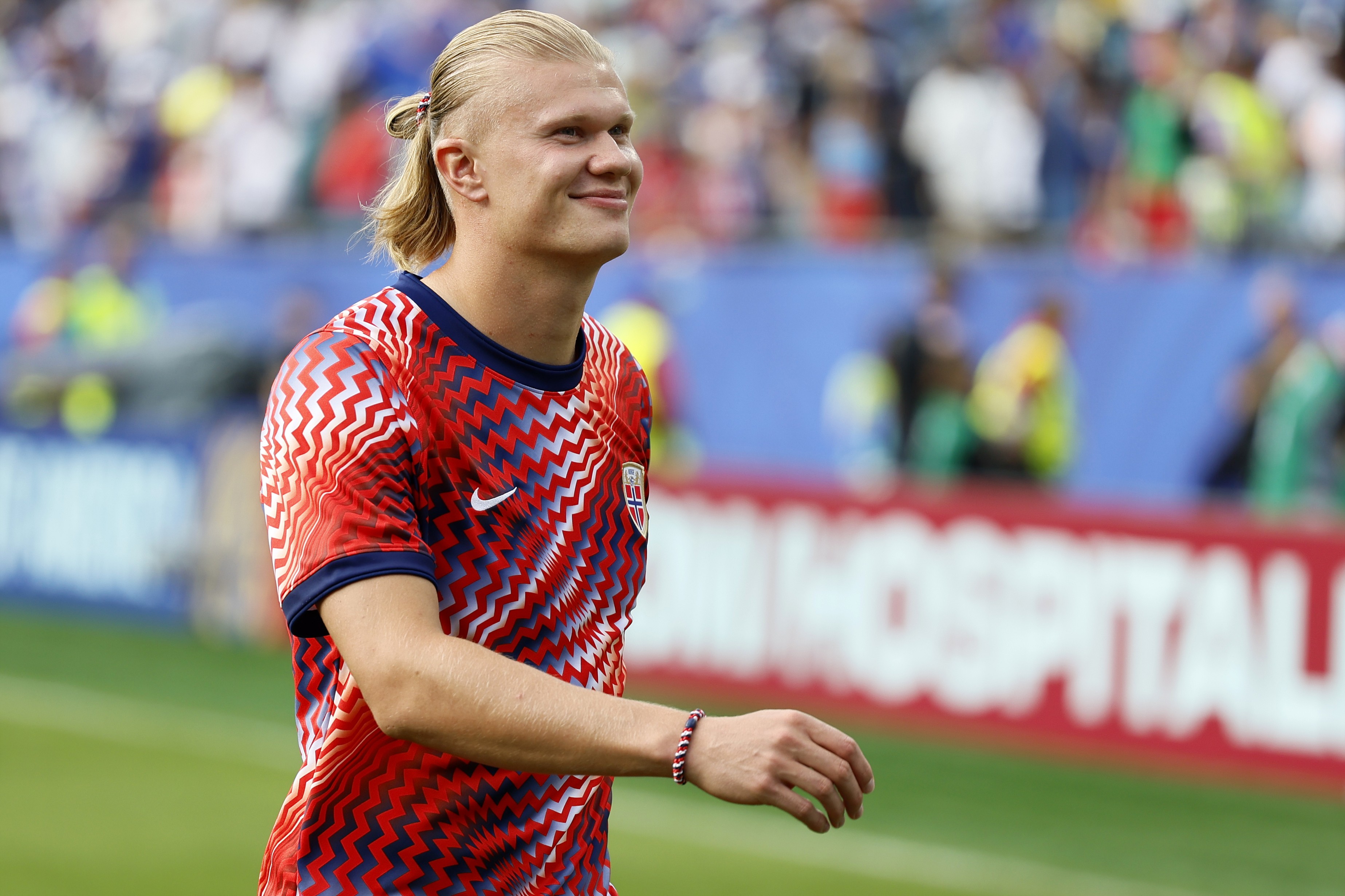 O jogador de futebol Erling Haaland, loiro de cabelo preso, sorri levemente enquanto veste uma camisa vermelha e azul com estampa em zigue-zague, em um campo de futebol com torcedores desfocados ao fundo