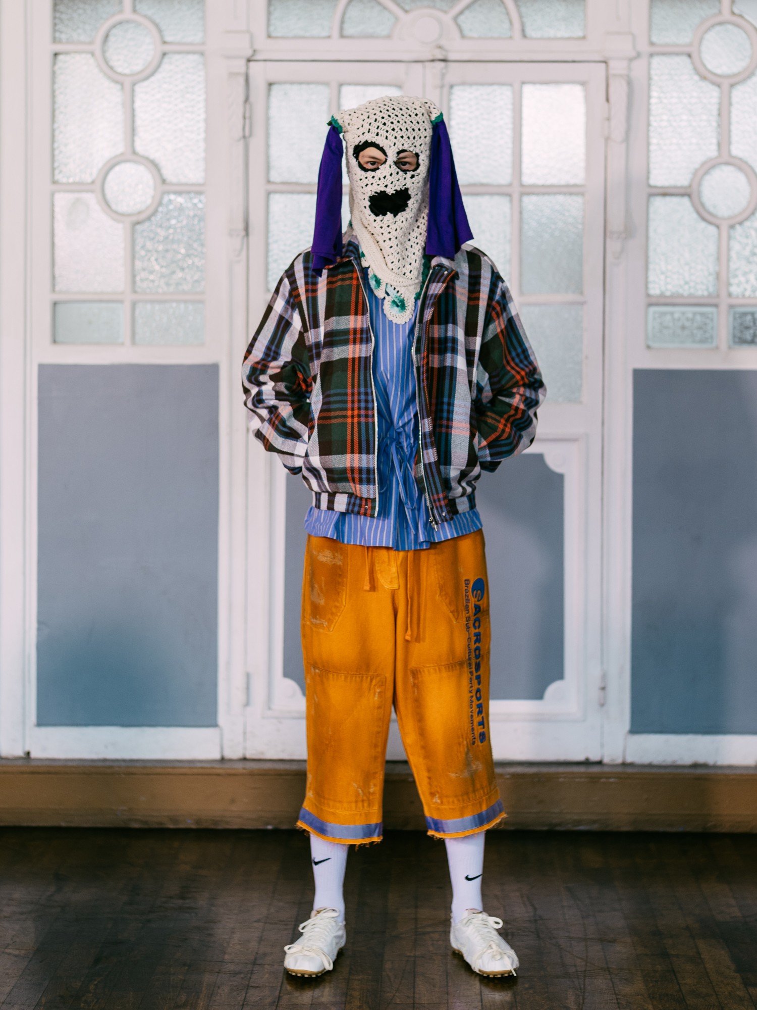 Pessoa com balaclava de crochê bege, olhos e boca pretos, e tecido roxo nas laterais. Veste jaqueta xadrez, camisa listrada azul e branca, bermuda laranja com estampa azul e tênis brancos. Posa em frente a uma porta branca com vidros texturizados.