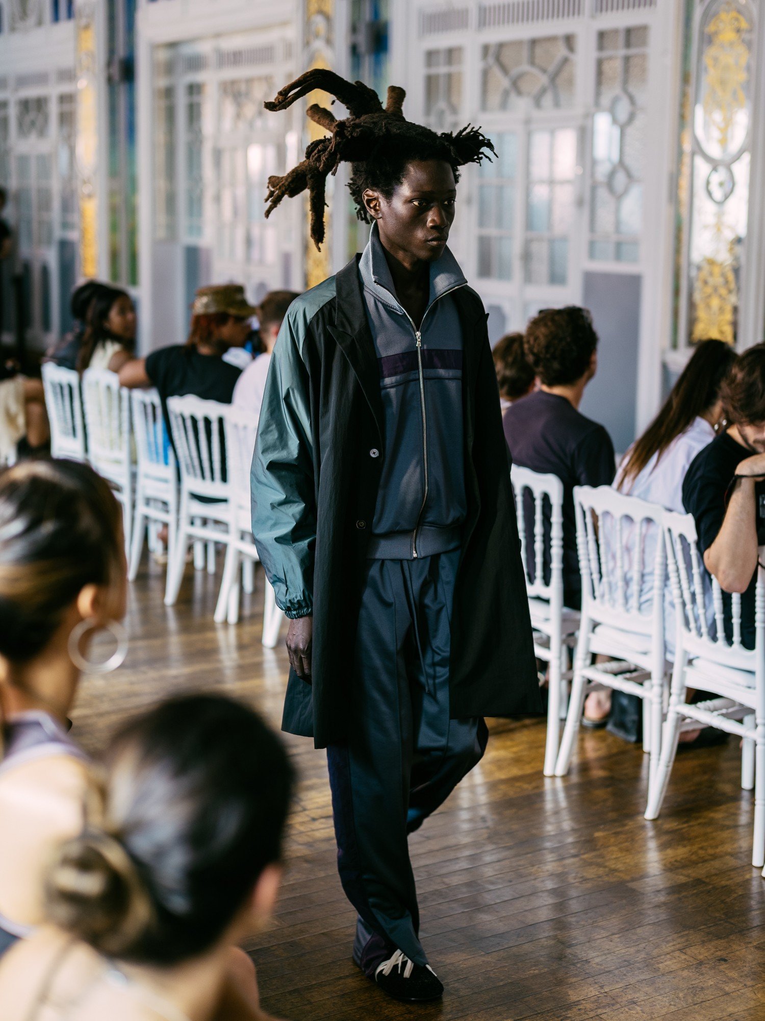 Modelo negro com dreadlocks espetados, vestindo casaco preto sobre agasalho cinza e calça escura, desfila em passarela de madeira. Ao fundo, pessoas sentadas em cadeiras brancas observam, em um salão com janelas altas e detalhes dourados