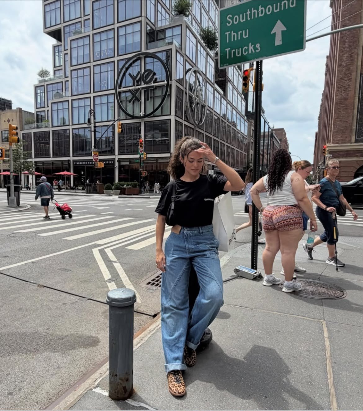 Jovem mulher de cabelo cacheado, blusa preta, calça jeans larga e sapatos de oncinha, com a mão na testa, em uma calçada movimentada de Nova York. Ao fundo, um prédio moderno com janelas grandes e um letreiro verde indicando Southbound Thru Trucks.