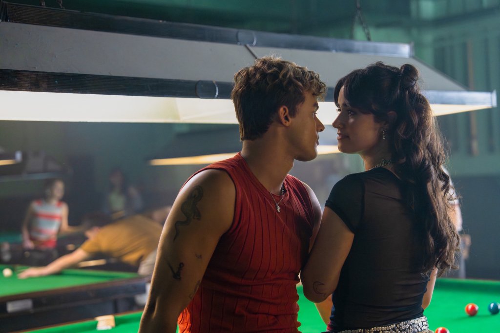 Um homem e uma mulher jovens se encaram de perfil em uma sinuca escura. Ele, à esquerda, usa regata vermelha e tem tatuagens no braço. Ela, à direita, tem cabelo escuro ondulado e usa blusa preta transparente. Ao fundo, outras pessoas jogam bilhar