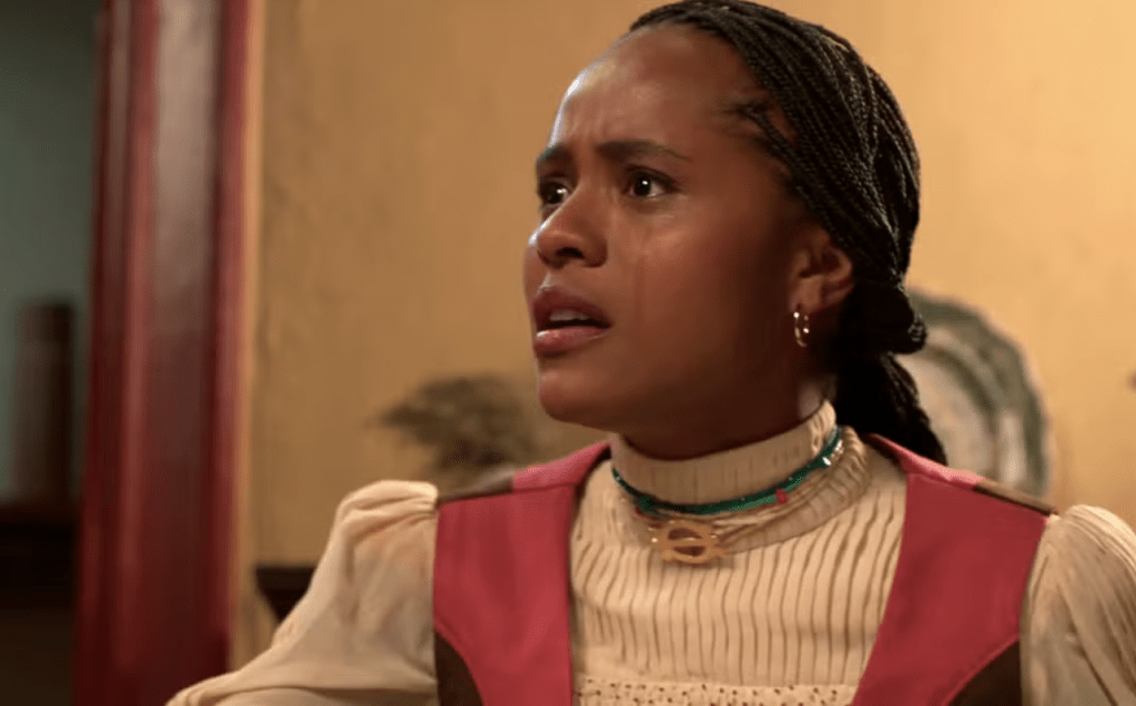Mulher negra com tranças, vestindo blusa creme e colete vermelho, olha para cima com expressão de preocupação e suor no rosto, em ambiente interno com parede bege