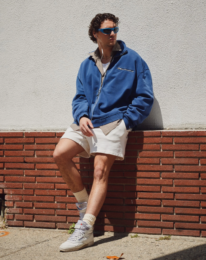 Homem jovem de cabelo cacheado escuro, óculos de sol azuis e fones de ouvido brancos, encostado em uma parede branca e tijolos vermelhos. Ele veste uma jaqueta azul, camisa xadrez, shorts creme e tênis brancos com meias altas