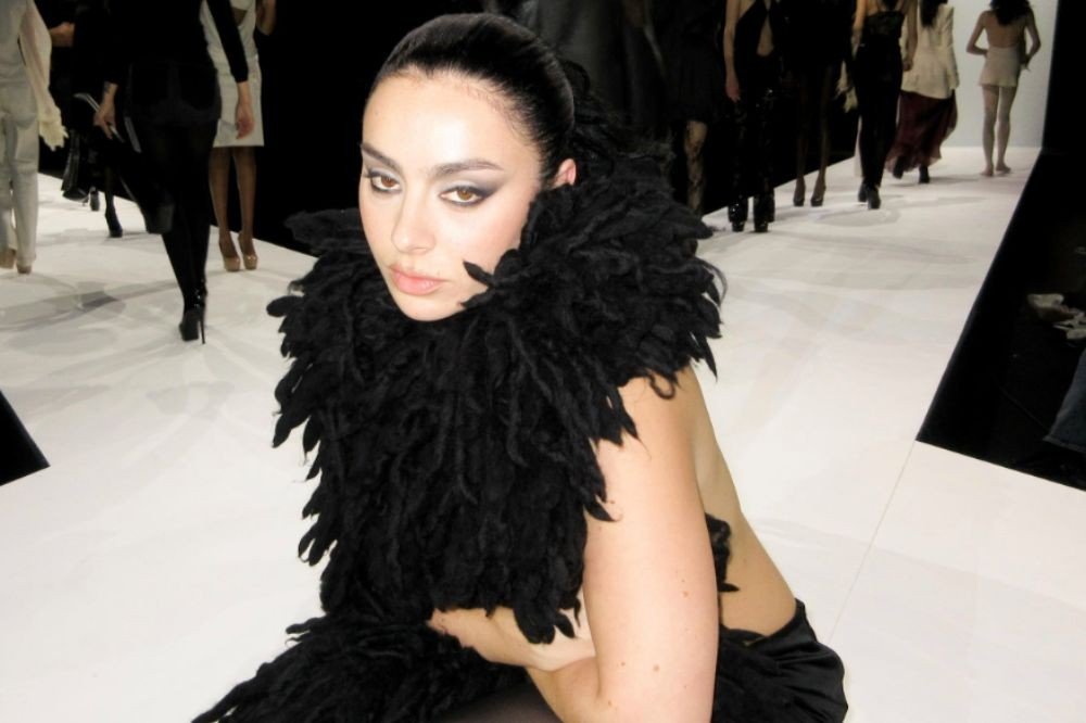 Mulher de cabelo escuro preso, maquiagem forte nos olhos, usando um adereço preto volumoso no pescoço e ombros, posa em uma passarela branca com detalhes pretos, enquanto modelos desfilam ao fundo
