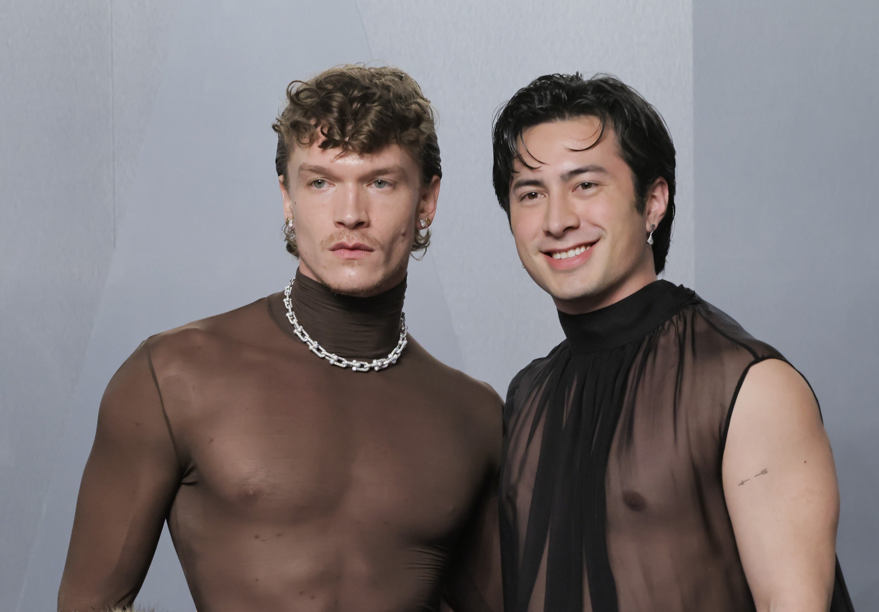 Dois homens posam lado a lado. O da esquerda, com cabelo encaracolado e bigode, veste blusa marrom transparente e colar prateado. O da direita, com cabelo liso escuro, sorri e usa blusa preta transparente sem mangas