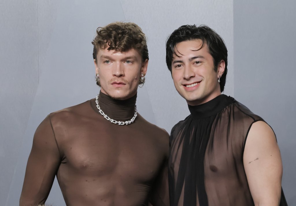 Dois homens posam lado a lado. O da esquerda, com cabelo encaracolado e bigode, veste blusa marrom transparente e colar prateado. O da direita, com cabelo liso escuro, sorri e usa blusa preta transparente sem mangas