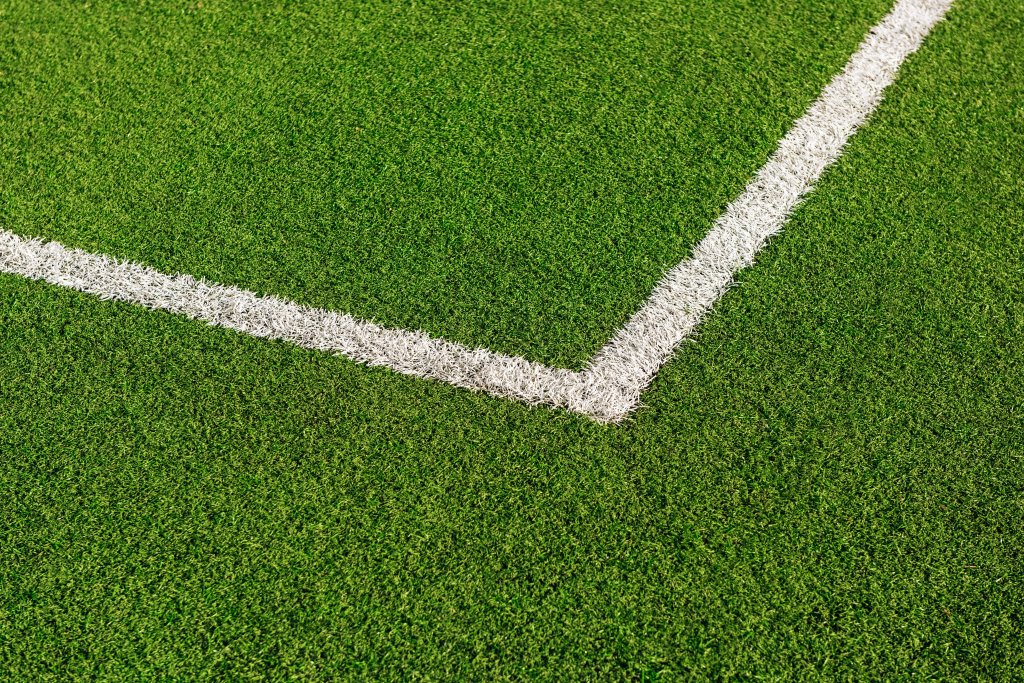 Campo de futebol com gramado sintético verde vibrante e duas linhas brancas marcando um canto do campo