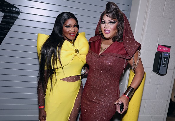 Duas drag queens sorrindo para a câmera. Uma veste um vestido amarelo com laço grande e recortes na cintura, a outra um vestido vinho brilhante com capuz e correntes no ombro. Ambas com maquiagem elaborada e cabelos longos.
