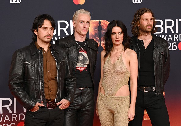 Quatro pessoas, três homens e uma mulher, posam no tapete vermelho do Brit Awards. Os homens vestem jaquetas de couro escuras, e a mulher usa um conjunto dourado de malha transparente