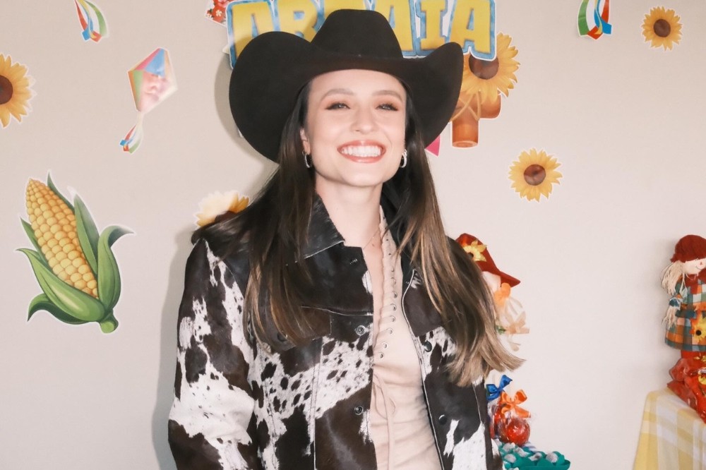Mulher jovem sorridente, de cabelos castanhos longos, usando chapéu de cowboy preto e jaqueta com estampa de vaca, em festa junina com decorações de milho, girassóis e balões coloridos