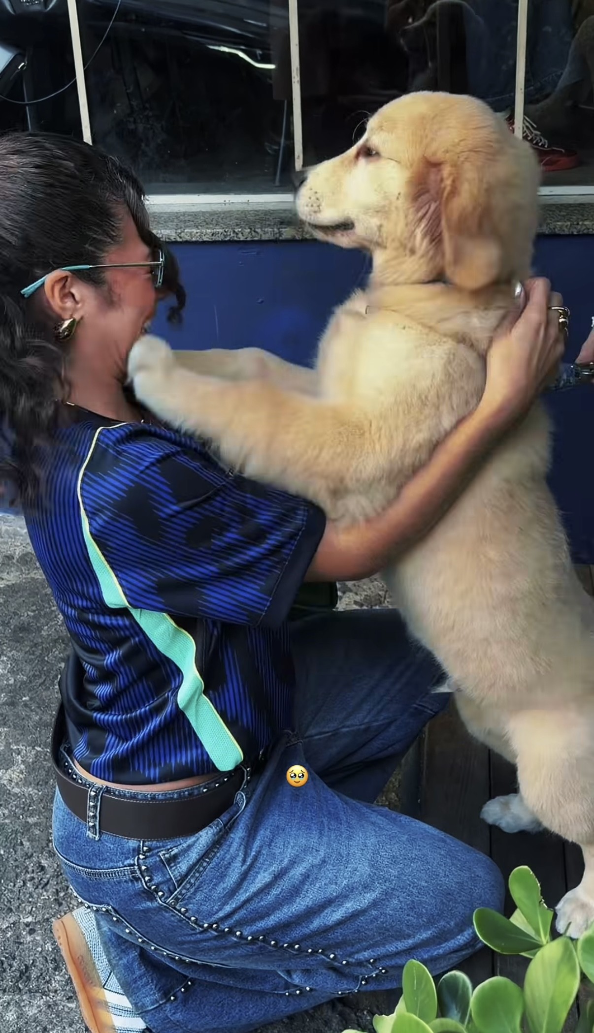 Mulher de cabelo escuro e óculos, ajoelhada, abraça um filhote de golden retriever que a lambe, ambos sorrindo. Ela veste camiseta azul com detalhes verdes e calça jeans com tachas, em um ambiente externo com plantas