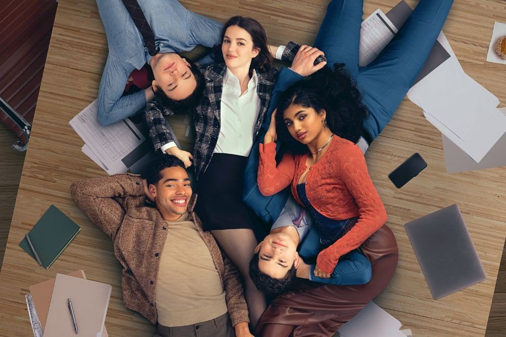 Cinco jovens, três homens e duas mulheres, deitados no chão de madeira clara, com papéis, cadernos e um notebook espalhados ao redor. Todos sorriem, exceto a mulher de blusa branca e blazer xadrez, que olha diretamente para a câmera. Um homem de camisa azul e gravata está no topo, e outro de jaqueta marrom sorri com as mãos atrás da cabeça. Uma mulher de blusa laranja e um homem de camisa cinza estão na parte inferior, com outro homem de terno azul no meio