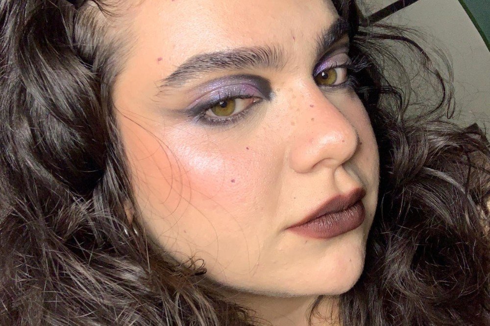 Rosto de mulher com cabelo cacheado escuro, maquiagem roxa nos olhos, delineado preto, batom marrom escuro e sardas no nariz