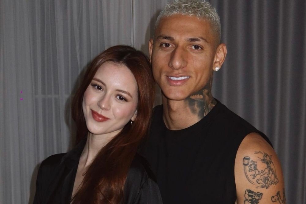 Mulher de cabelos ruivos e homem de cabelo loiro platinado, ambos sorrindo para a câmera. Ele veste regata preta e tem tatuagens no pescoço e braço. Ela usa batom terracota e blazer preto.