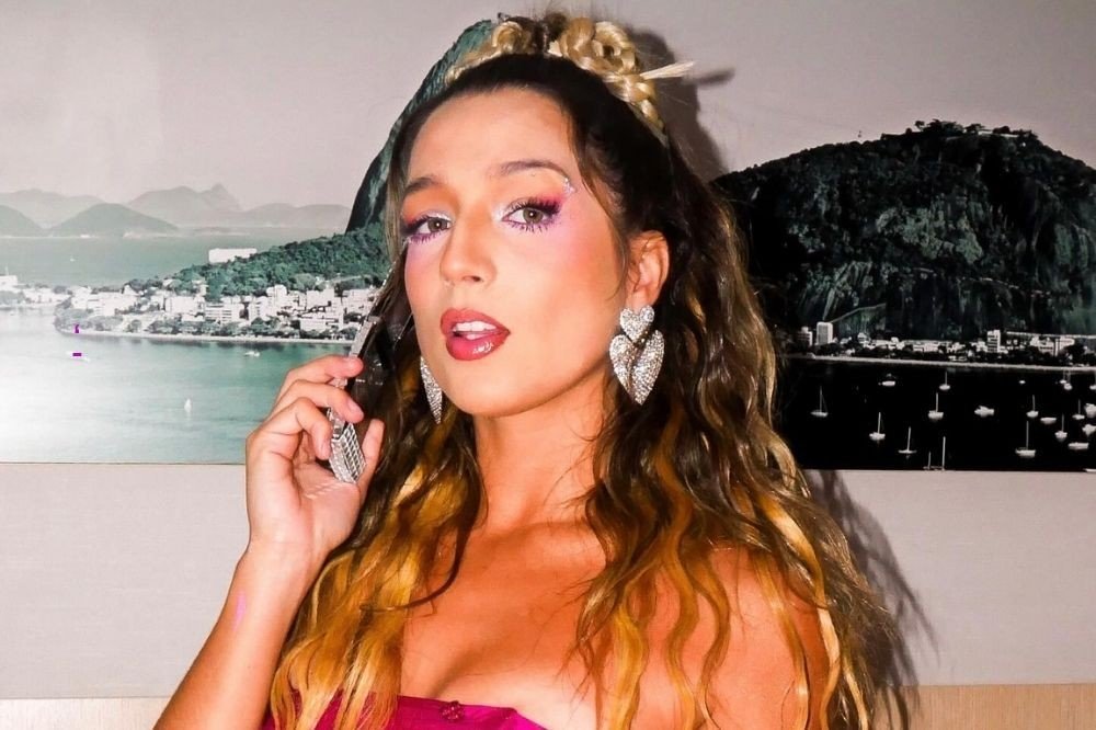 Mulher jovem com cabelo castanho ondulado e mechas loiras, maquiagem vibrante com sombra rosa e roxa, batom rosa. Ela usa brincos grandes em formato de coração cravejados e um top tomara que caia magenta. Ao fundo, quadros com paisagens do Rio de Janeiro, incluindo o Pão de Açúcar e a Baía de Guanabara