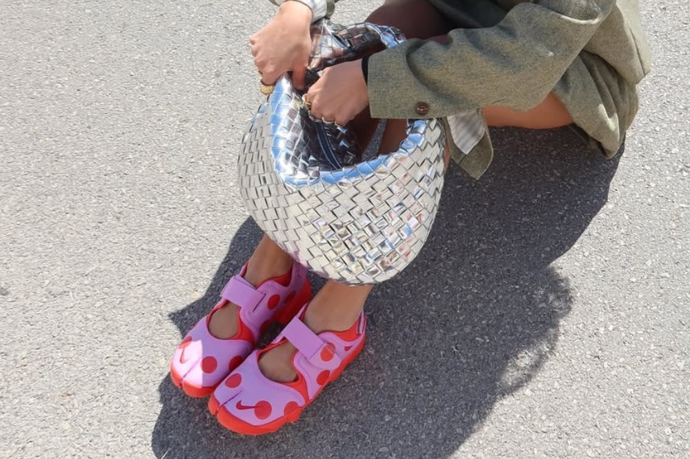 Pessoa sentada no chão de asfalto, usando sapatos rosa e lilás com bolinhas e uma bolsa prateada trançada. Veste blazer verde-oliva e segura a bolsa com as duas mãos, abrindo-a
