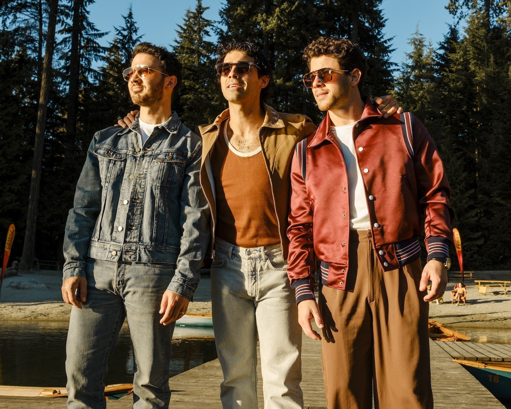 Três homens jovens, com óculos de sol, posam em um píer de madeira, com floresta e lago ao fundo. O da esquerda veste jaqueta jeans e calça cinza, o do meio usa jaqueta marrom e calça clara, e o da direita, jaqueta de cetim bordô e calça marrom. Todos olham para a direita, com expressão séria
