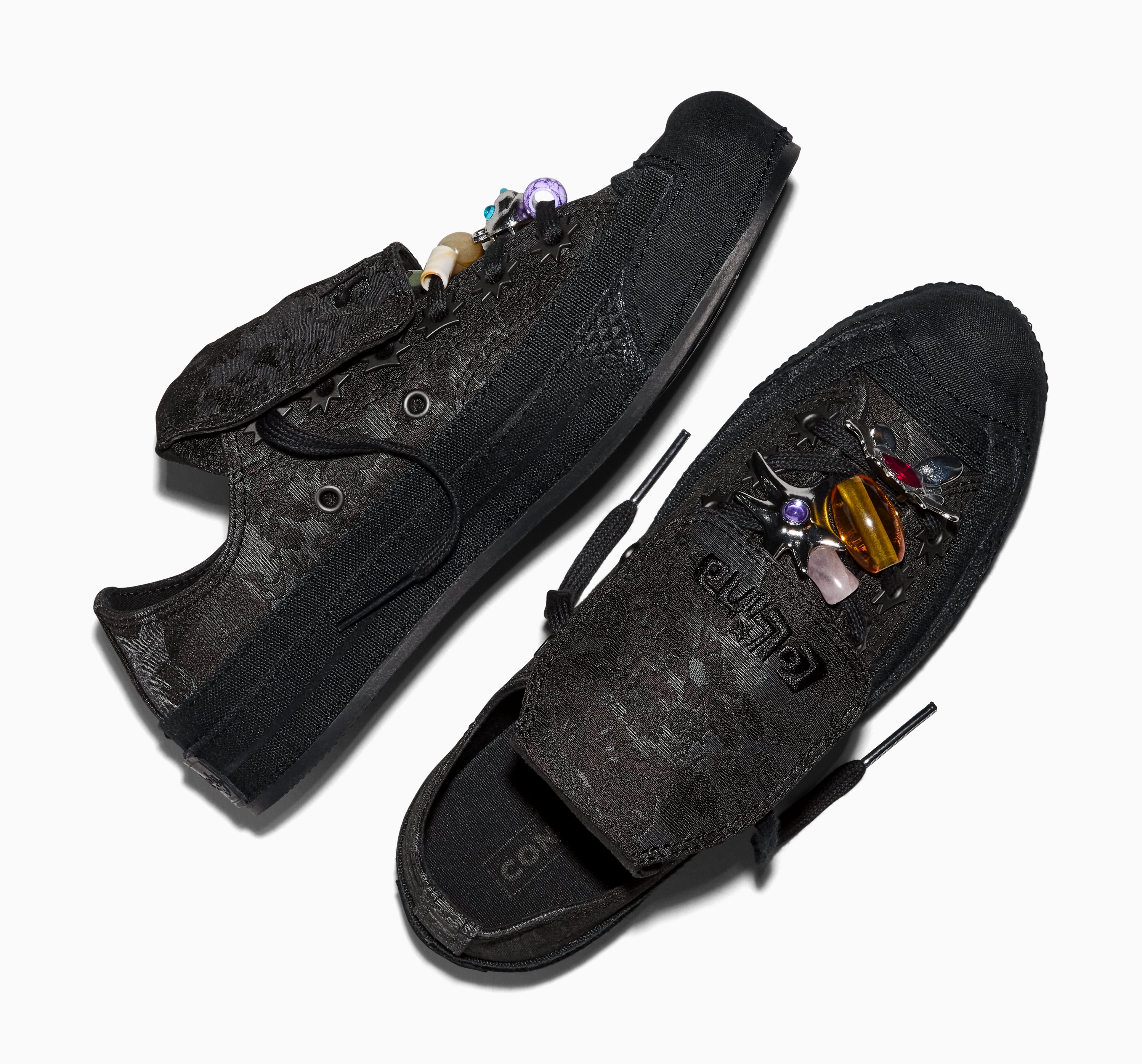 Par de tênis Converse pretos com estampa floral em relevo, um com pingentes de contas coloridas nos cadarços e outro com broches de insetos e pedras preciosas na lingueta