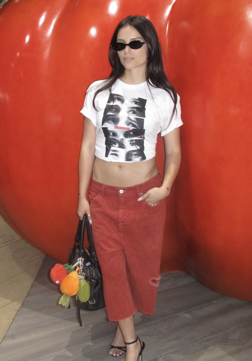 Mulher de cabelos longos e escuros, óculos de sol ovais, camiseta branca cropped com estampa de olhos e a palavra OBSESSION, calça jeans vermelha rasgada no joelho, sandálias de salto alto pretas e bolsa preta com chaveiros de frutas, posando em frente a uma parede vermelha brilhante
