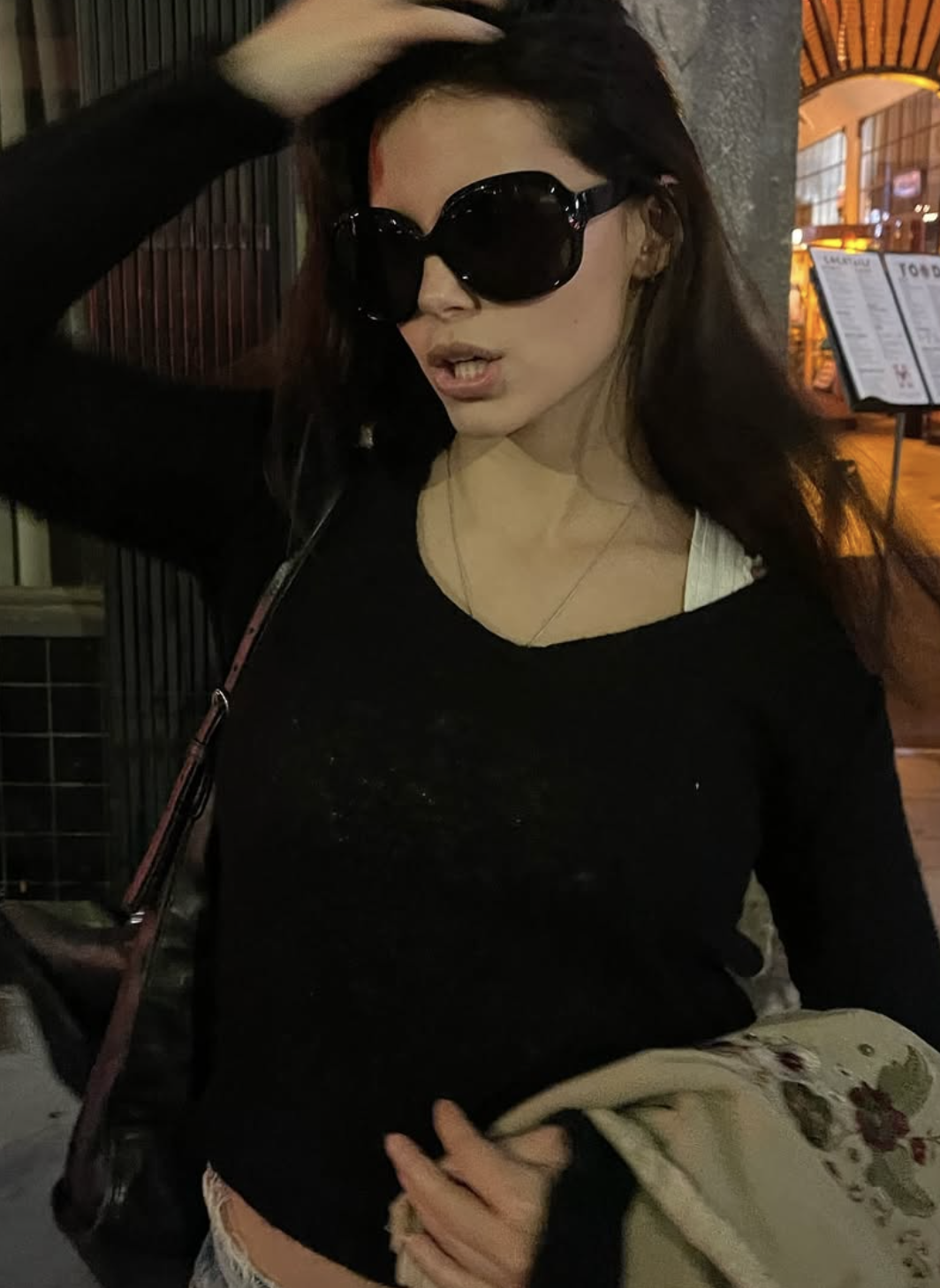 Jovem mulher com cabelos longos e escuros, óculos de sol grandes, blusa preta e colar fino, ajustando o cabelo com a mão direita. Ela tem a boca entreaberta, segurando um casaco estampado na mão esquerda, em um ambiente noturno urbano com luzes ao fundo