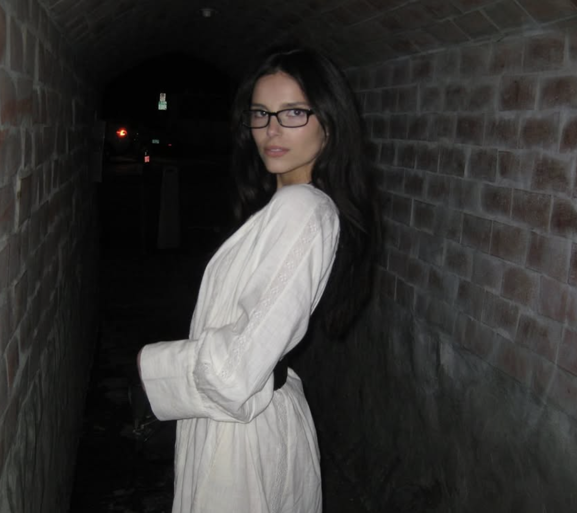 Mulher de cabelos longos e escuros, usando óculos e um vestido branco, posa de lado em um túnel escuro de tijolos, olhando para a câmera