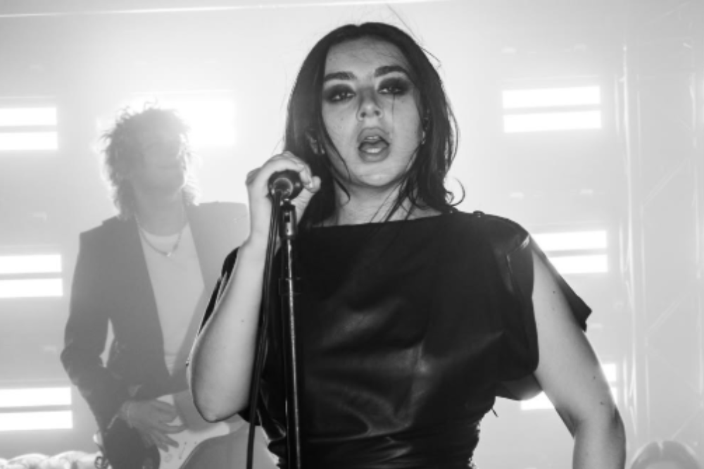 Cantora de cabelo escuro, maquiagem pesada e blusa preta de couro, cantando em um microfone com a boca aberta, em um palco iluminado. Um guitarrista de cabelo encaracolado e terno escuro aparece desfocado ao fundo