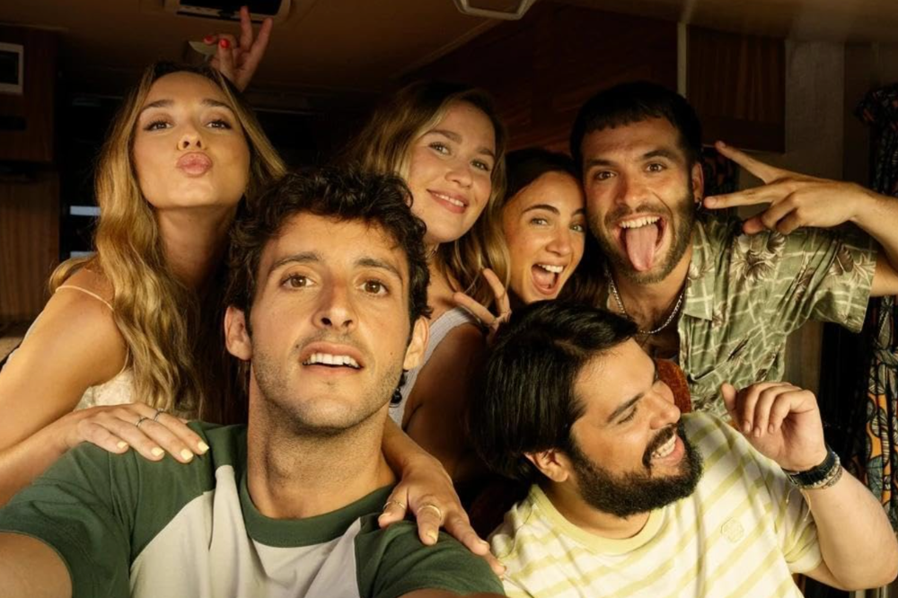 Seis jovens adultos, três homens e três mulheres, tiram uma selfie divertida. Um homem na frente, com camiseta verde e branca, olha para cima. Atrás dele, uma mulher loira faz bico e sinal de paz, outra sorri, e um homem barbudo mostra a língua e faz sinal de rock. Outra mulher e um homem barbudo riem ao lado, todos com expressões alegres