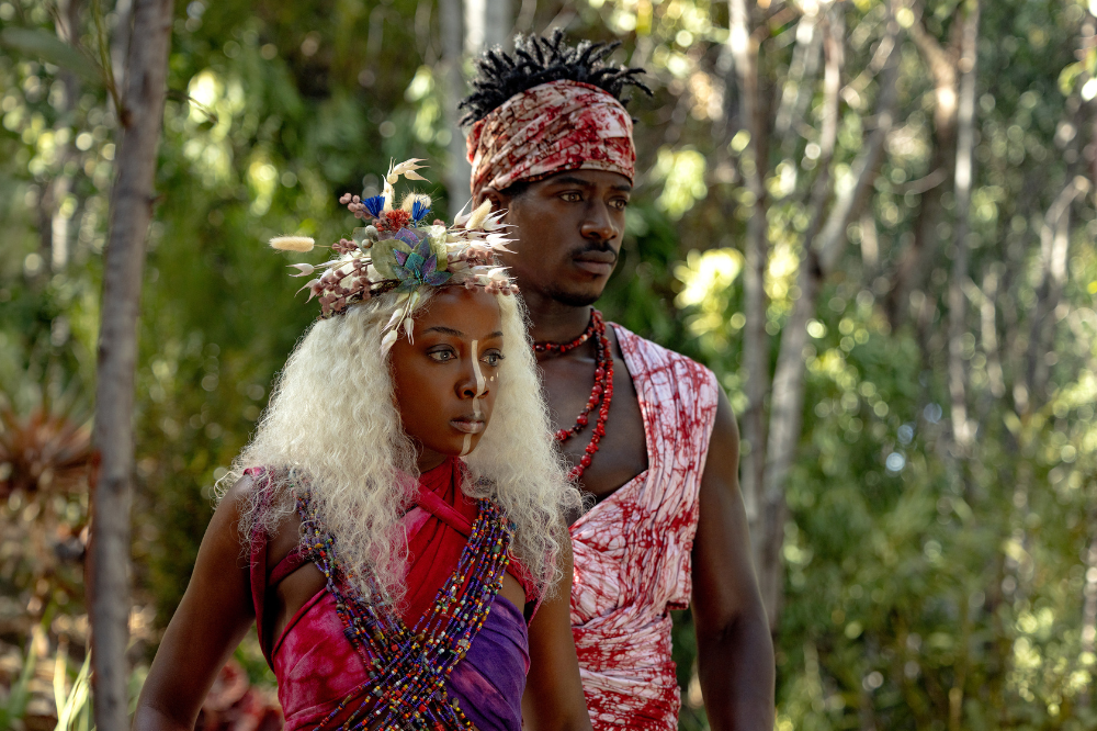 Uma mulher e um homem negros em trajes cerimoniais, em meio a uma floresta. A mulher, à frente, tem cabelos loiros cacheados, maquiagem facial branca e um adorno floral na cabeça. Veste um top colorido de miçangas e tecido. O homem, atrás, usa um turbante e uma túnica vermelha e branca, com um colar de contas vermelhas. Ambos olham para a direita, com expressões sérias