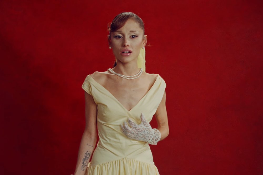 Cantora Ariana Grande com expressão de surpresa, usando um vestido creme estilo vintage, colar de pérolas e luvas de renda brancas, em fundo vermelho