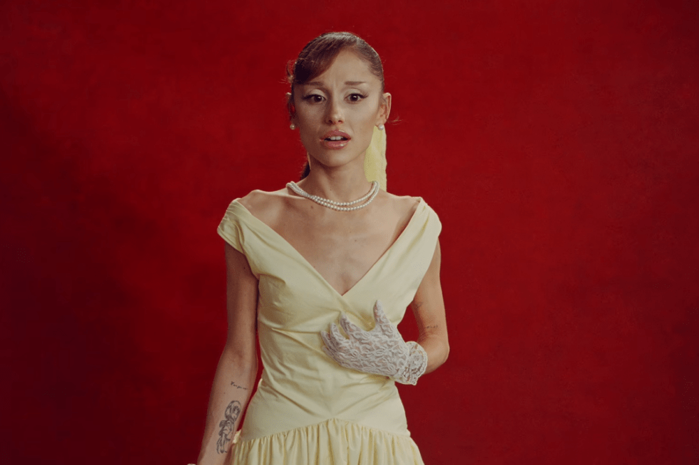 Cantora Ariana Grande com expressão de surpresa, usando um vestido creme estilo vintage, colar de pérolas e luvas de renda brancas, em fundo vermelho