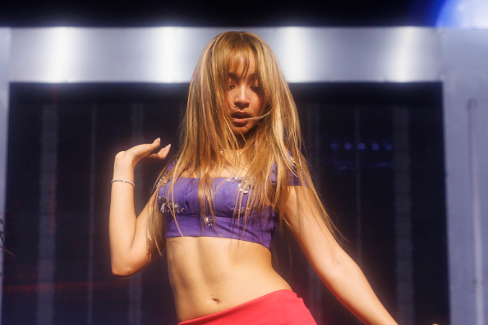 Mulher de cabelos longos e loiros dançando, vestindo um top roxo brilhante e calça vermelha, com a boca entreaberta e os braços levantados, em um palco escuro com luzes azuis