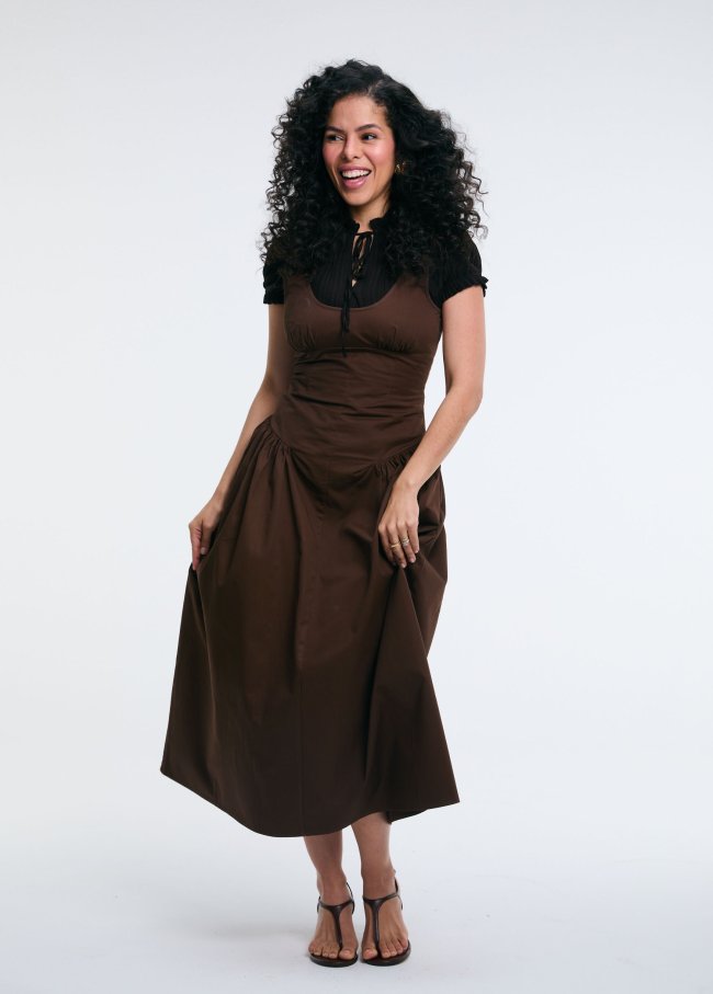 Mulher de pele morena e cabelos cacheados longos, sorrindo e olhando para a direita, vestindo um vestido marrom sobre uma blusa preta de mangas curtas, em fundo branco