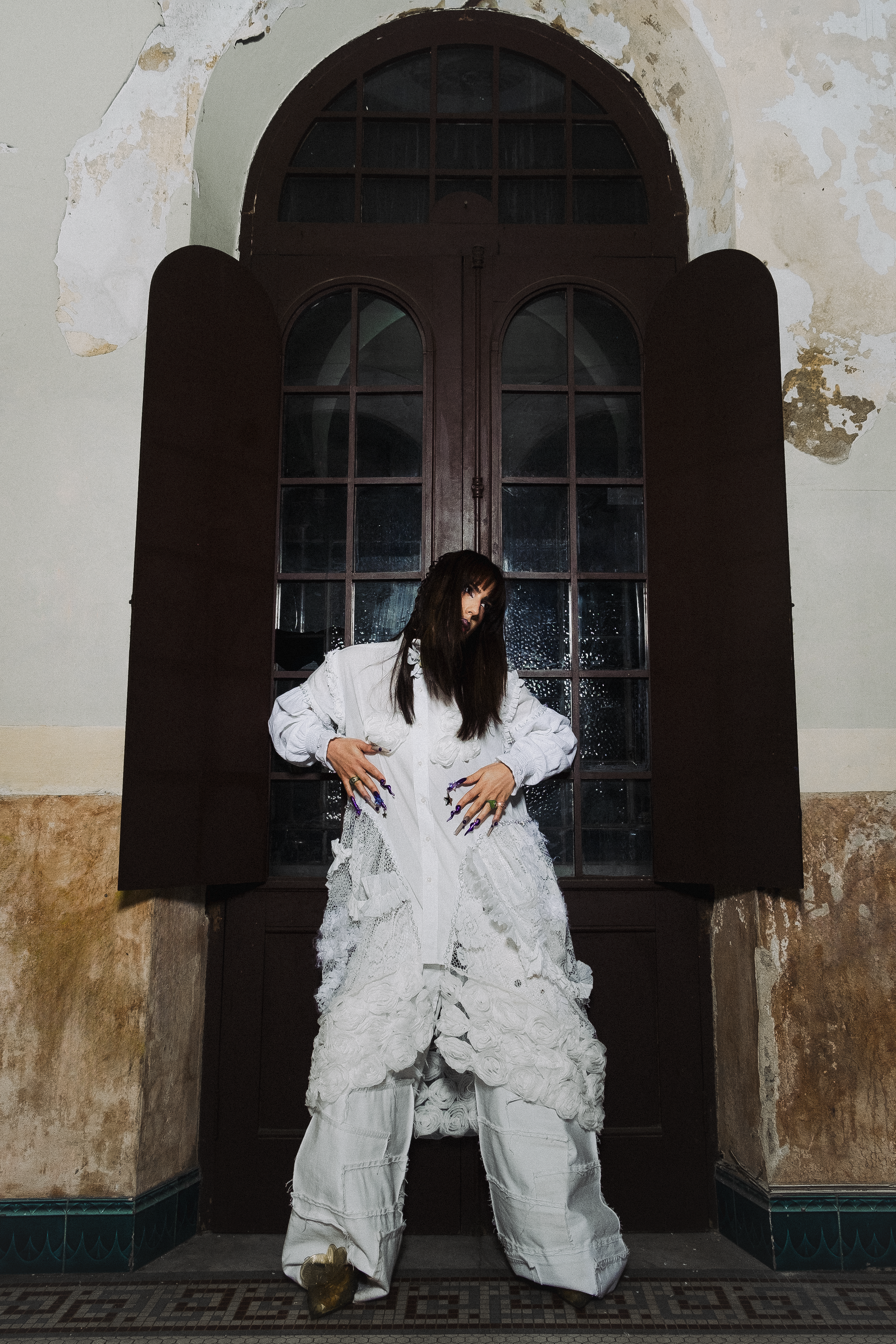 Pessoa com cabelo escuro e franja, vestindo camisa branca oversized e calças largas brancas com detalhes de renda, posa em frente a uma porta de madeira escura com janelas arqueadas. As paredes ao redor estão descascadas, revelando a textura antiga