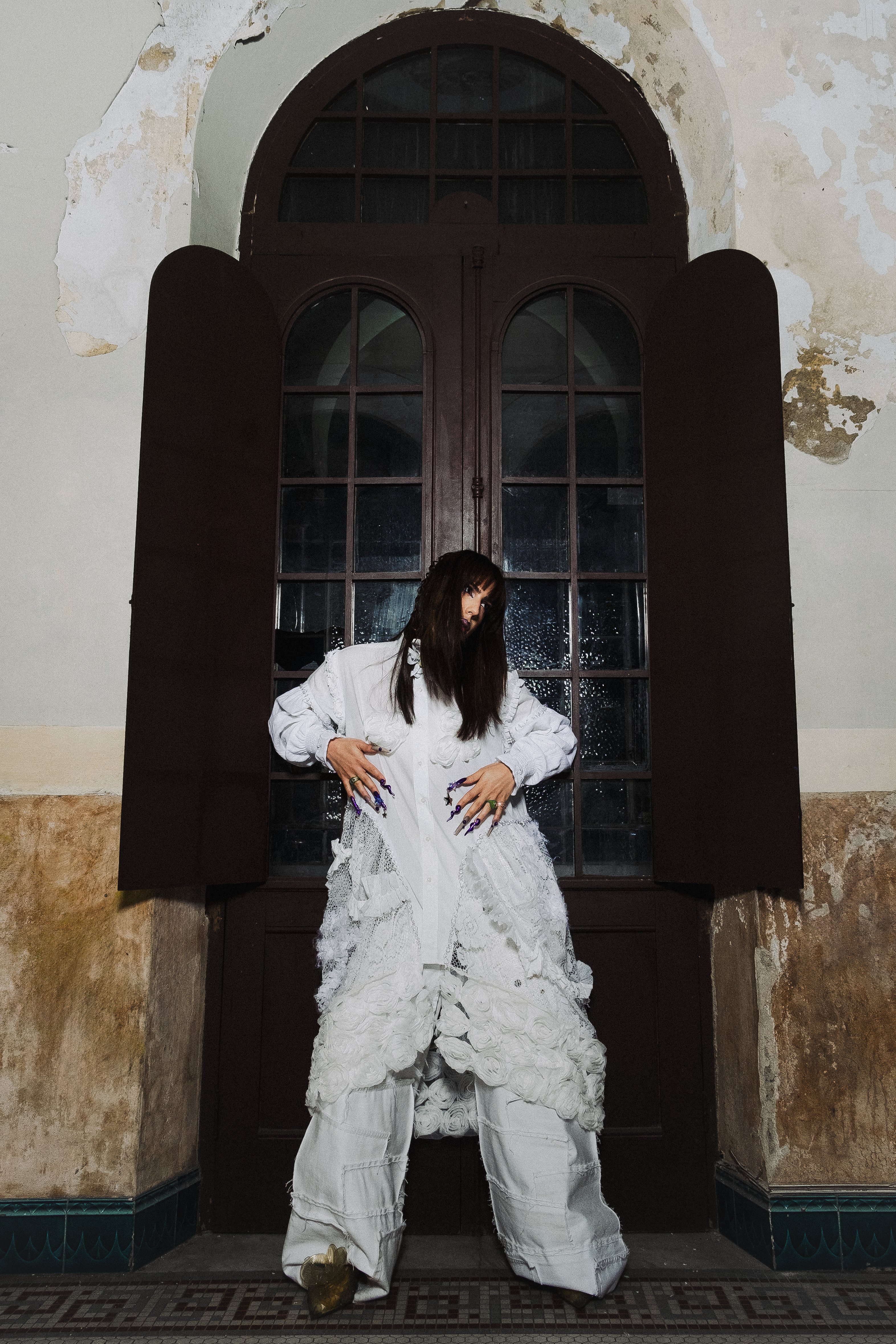 Pessoa com cabelo escuro e franja, vestindo camisa branca oversized e calças largas brancas com detalhes de renda, posa em frente a uma porta de madeira escura com janelas arqueadas. As paredes ao redor estão descascadas, revelando a textura antiga