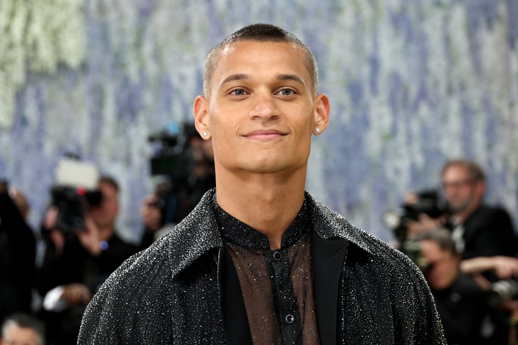 Homem jovem de pele morena e cabelo raspado, sorrindo levemente, usando brincos de ponto de luz e uma camisa preta brilhante sobre uma blusa transparente marrom, em um evento com fundo desfocado de flores azuis e pessoas